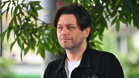 Gabriel Fleszar z ciemnymi, długimi włosami, w czarnej kurtce, stoi wśród zieleni.