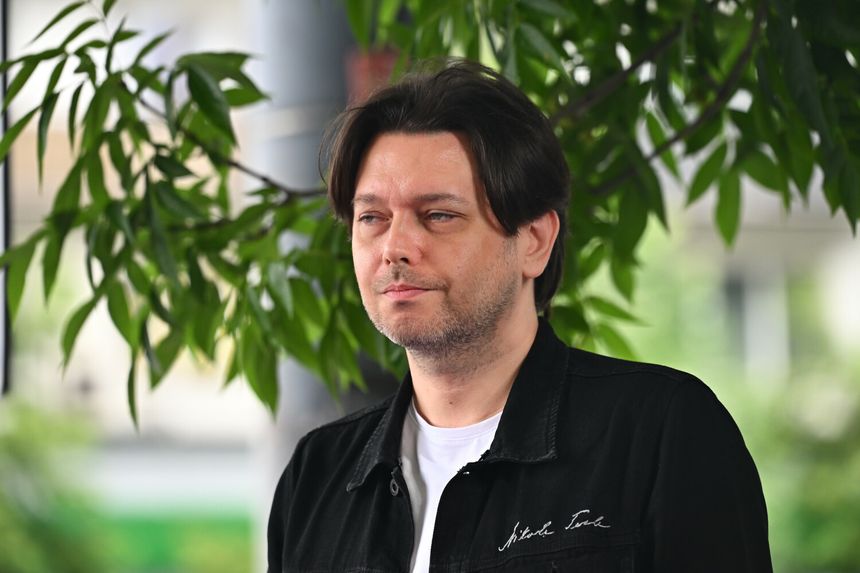 Gabriel Fleszar z ciemnymi, długimi włosami, w czarnej kurtce, stoi wśród zieleni.
