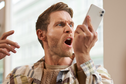 Mężczyzna w plaidowej koszuli, zdenerwowany, krzyczy w telefon. Tło jasne, okna za nim.