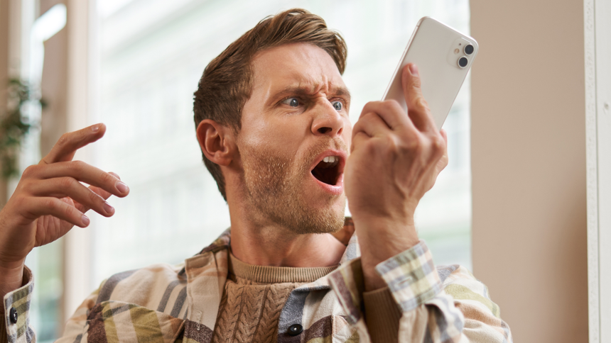 Mężczyzna w plaidowej koszuli, zdenerwowany, krzyczy w telefon. Tło jasne, okna za nim.
