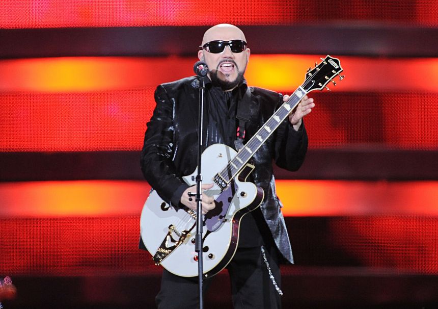 Mężczyzna w ciemnych okularach, z białą gitarą, śpiewa na tle czerwonych świateł.