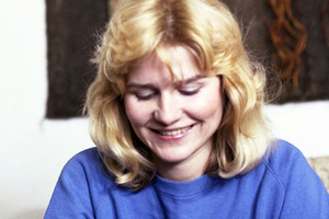 Na zdjęciu uśmiechnięta kobieta z blond włosami, ubrana w niebieską bluzę, skupiona na czymś.