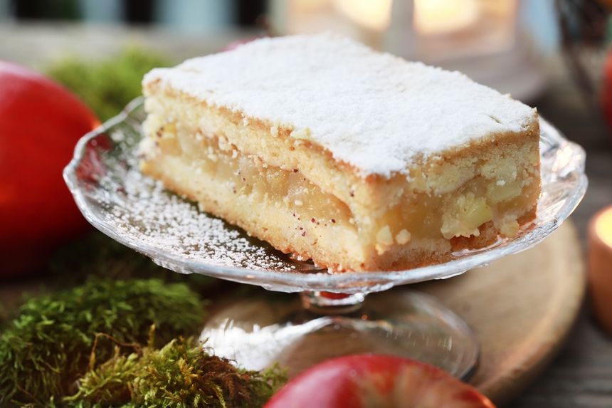 Ciasto z jabłkami, posypane cukrem pudrem, na szklanym talerzu, obok czerwone jabłka.