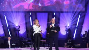 Dwoje prowadzących Taniec z gwiazdami w TVN na scenie, mikrofony w rękach, w tle efekty świetlne i zespół muzyczny.