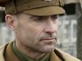 Mężczyzna w mundurze wojskowym z brązową czapką, patrzący w dal. Tło w odcieniach szarości.