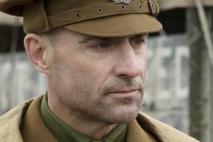 Mężczyzna w mundurze wojskowym z brązową czapką, patrzący w dal. Tło w odcieniach szarości.