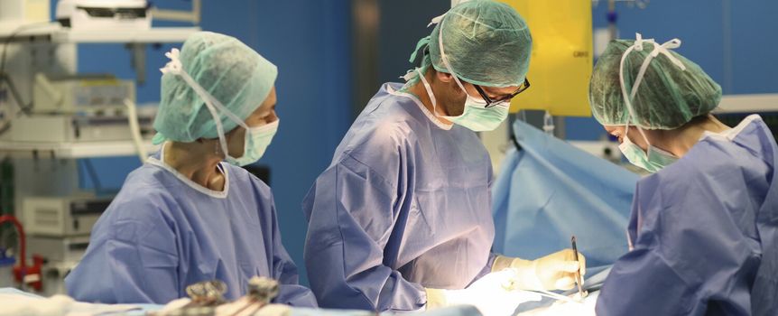 Grupa chirurgów w szpitalu skupiona na operacji, w maseczkach i odzieży ochronnej.