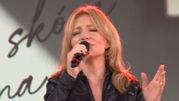 Kobieta z blond włosami, w czarnej kurtce, śpiewa do mikrofonu na scenie.