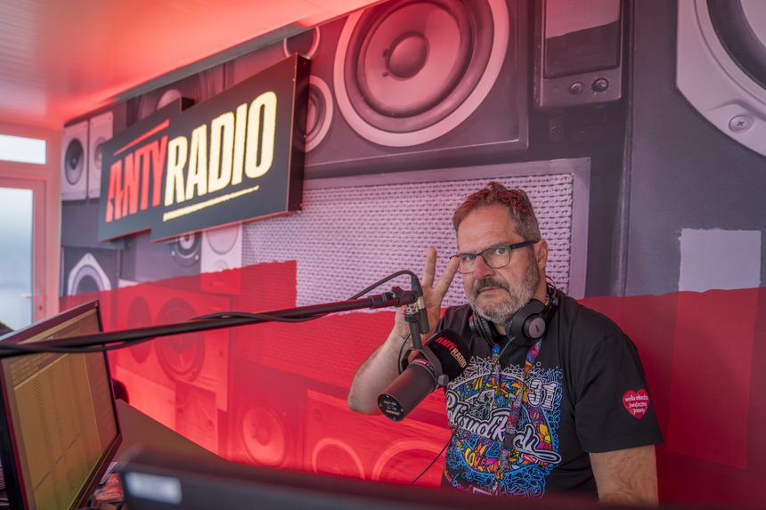 Mężczyzna w okularach, z mikrofonem, robi znak „pokoju” w studiu radiowym. Tło w czerwonym świetle.