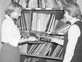 Dwie kobiety w bibliotece wymieniają się płytami, otaczają je półki z albumami.