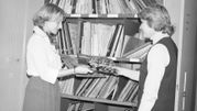 Dwie kobiety w bibliotece wymieniają się płytami, otaczają je półki z albumami.