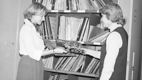 Dwie kobiety w bibliotece wymieniają się płytami, otaczają je półki z albumami.
