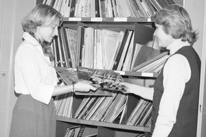 Dwie kobiety w bibliotece wymieniają się płytami, otaczają je półki z albumami.