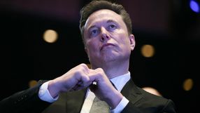 Na zdjęciu mężczyzna w garniturze z gestem dłoni, w tle rozmyte światła.