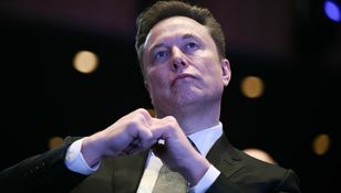 Na zdjęciu mężczyzna w garniturze z gestem dłoni, w tle rozmyte światła.