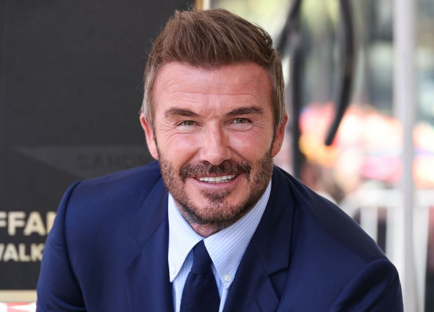 David Beckham w niebieskim garniturze, uśmiechnięty, z krótkimi, stylowymi włosami.