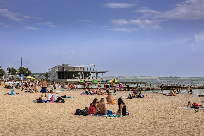 Plaża z wczasowiczami na piasku, budynek w tle, woda w pobliżu. Słoneczny dzień.