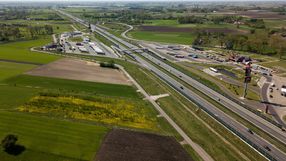 Widok z lotu ptaka na rozległe tereny zielone z drogą oraz zabudowaniami przy trasie.