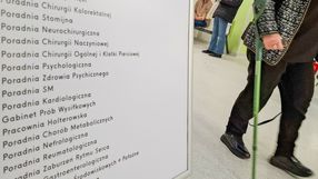 Zdjęcie przedstawia pacjenta idącego w kierunku tablicy z nazwami poradni medycznych.