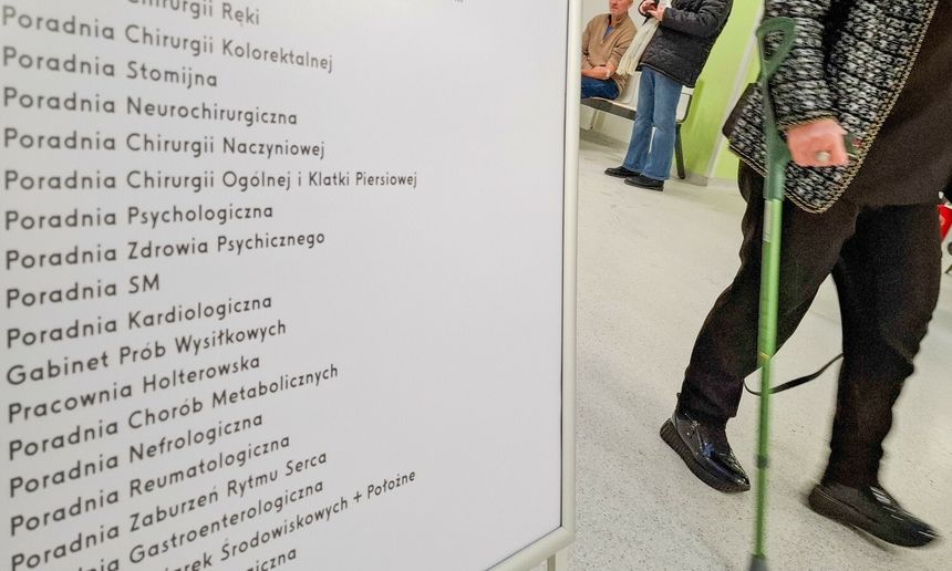 Zdjęcie przedstawia pacjenta idącego w kierunku tablicy z nazwami poradni medycznych.