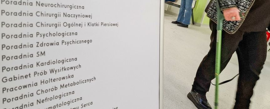 Zdjęcie przedstawia pacjenta idącego w kierunku tablicy z nazwami poradni medycznych.