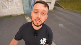 Książulo w czarnej koszulce, na tle szarego muru, mówi do kamery przy ulicy.