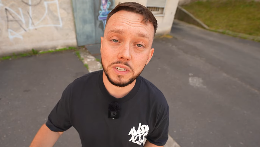 Książulo w czarnej koszulce, na tle szarego muru, mówi do kamery przy ulicy.