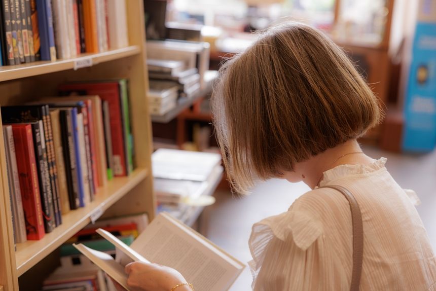 Kobieta z krótkimi włosami przegląda książki w bibliotece. W tle regały z książkami.