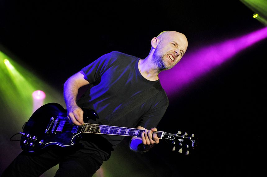 Muzyk gra na gitarze elektrycznej, na tle kolorowych świateł scenicznych.