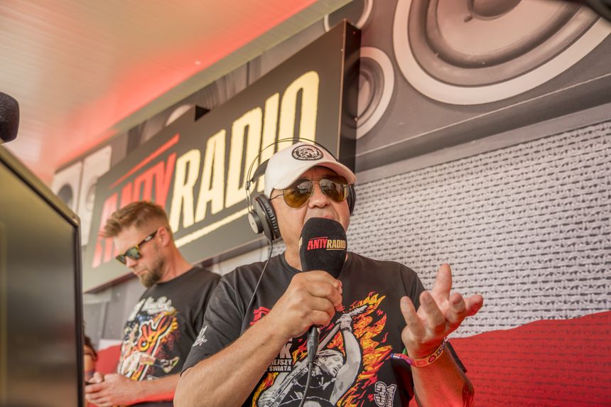 Mężczyzna w ciemnych okularach i słuchawkach mówi do mikrofonu na tle logo antyRADIO.