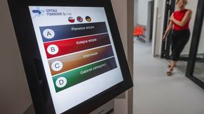 Wyświetlacz dotykowy z menu informacyjnym w nowoczesnym szpitalu. W tle przechodzi osoba.