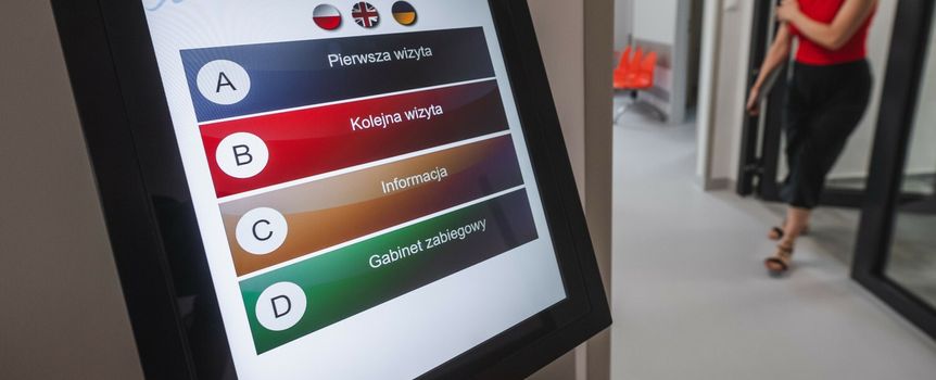 Wyświetlacz dotykowy z menu informacyjnym w nowoczesnym szpitalu. W tle przechodzi osoba.