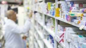 Apteka, na półkach różne leki, farmaceuta w białym fartuchu sięga po produkt.
