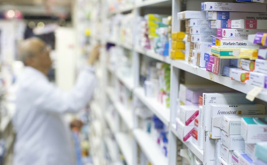 Apteka, na półkach różne leki, farmaceuta w białym fartuchu sięga po produkt.