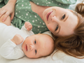 Rudowłosa mama w zielonej sukience w groszki leżąca na łóżku z dzieckiem w białych śpioszkach