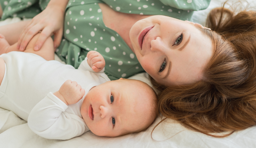 Rudowłosa mama w zielonej sukience w groszki leżąca na łóżku z dzieckiem w białych śpioszkach