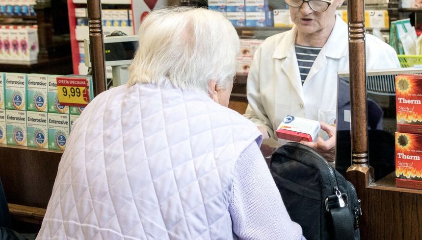 Kobieta w jasnej kurtce rozmawia z farmaceutką w aptece. W tle półki z lekami.