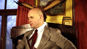 Mężczyzna w garniturze, siedzący na krześle z orłem w tle, na czerwonym tle.