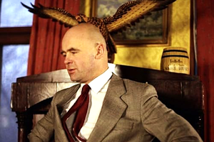 Mężczyzna w garniturze, siedzący na krześle z orłem w tle, na czerwonym tle.