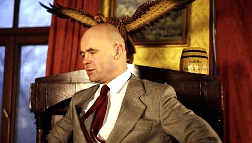 Mężczyzna w garniturze, siedzący na krześle z orłem w tle, na czerwonym tle.