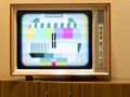 Telewizor retro z testowym obrazem; kolorowe paski i napisy. Stoi na drewnianym meblu.