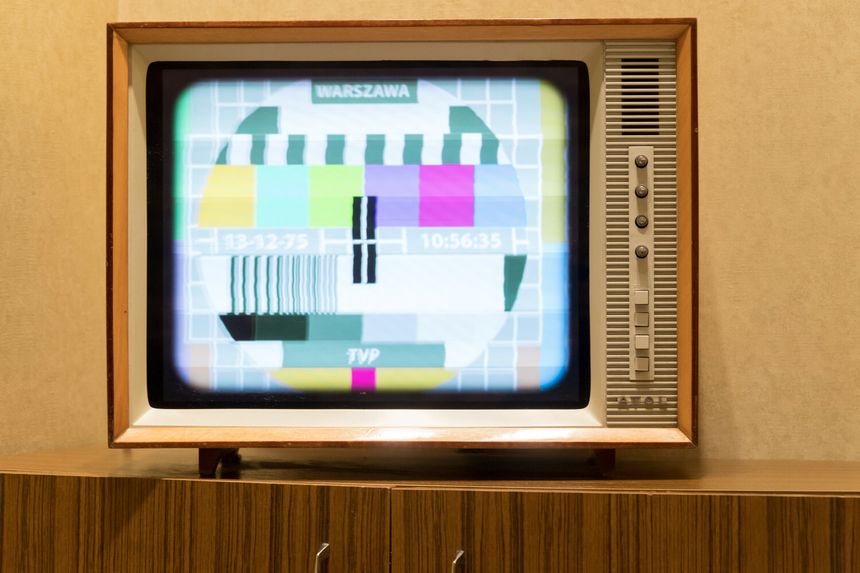 Telewizor retro z testowym obrazem; kolorowe paski i napisy. Stoi na drewnianym meblu.