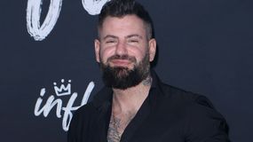 Wojtek Gola z brodą w czarnej koszuli, uśmiecha się na tle z napisem 