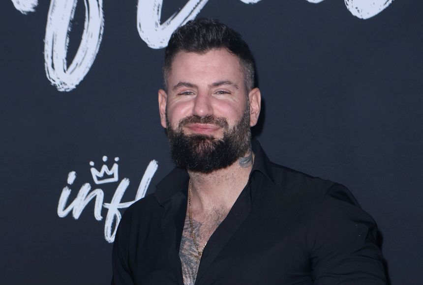 Wojtek Gola z brodą w czarnej koszuli, uśmiecha się na tle z napisem 