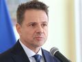 Mężczyzna w garniturze mówi do mikrofonu, w tle widoczne flagi. Wyraz twarzy poważny.