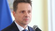 Mężczyzna w garniturze mówi do mikrofonu, w tle widoczne flagi. Wyraz twarzy poważny.