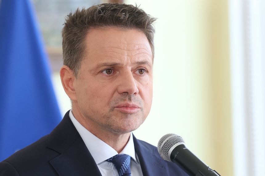 Mężczyzna w garniturze mówi do mikrofonu, w tle widoczne flagi. Wyraz twarzy poważny.