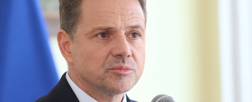 Mężczyzna w garniturze mówi do mikrofonu, w tle widoczne flagi. Wyraz twarzy poważny.