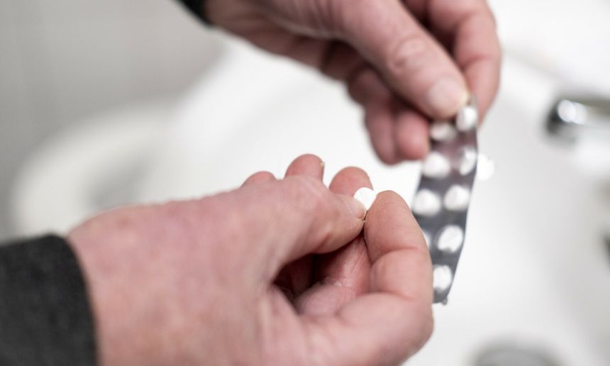 Osoba trzyma w dłoni tabletkę i opakowanie leków w łazience.