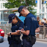 Dwoje policjantów na ulicy, sprawdzają dokumenty kierowcy w samochodzie. W tle przechodnie.